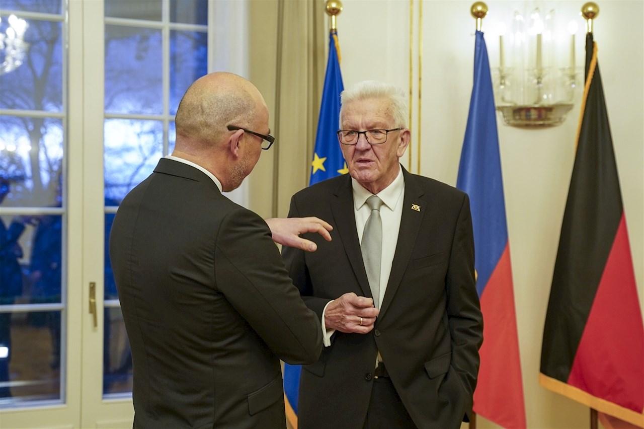 Regierungschef Daniel Risch spricht mit Baden-Württembergs Ministerpräsident Winfried Kretschmann. Im Hintergrund die Flaggen der EU, Liechtensteins und Deutschlands sowie eine Fensterfront.